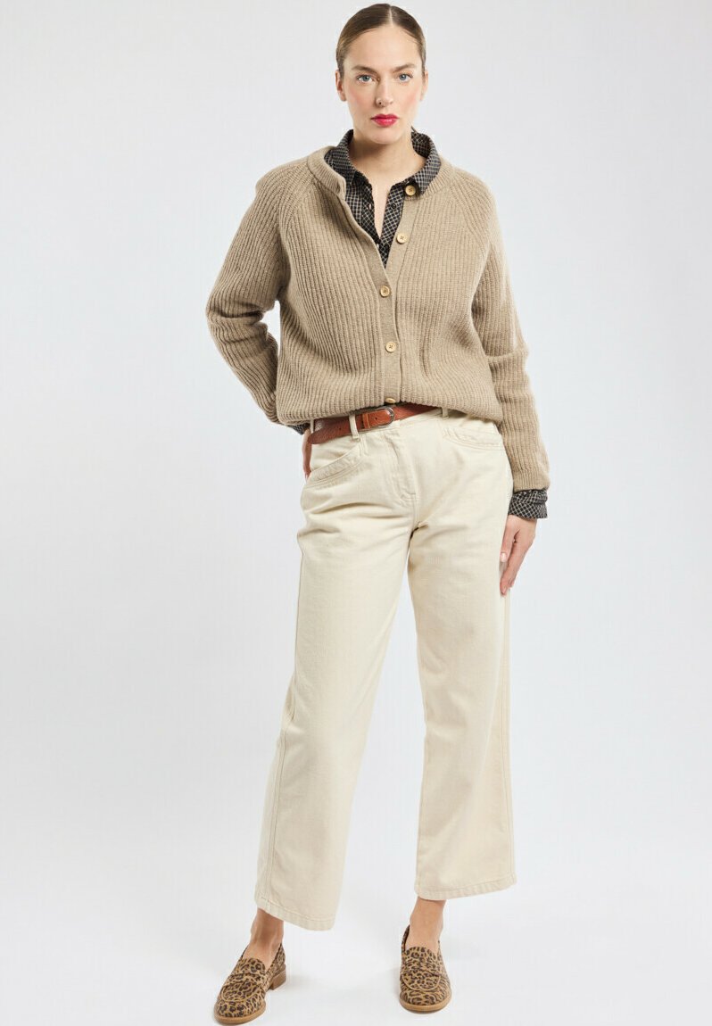 Beige geribbeld vest met houten knopen, over een zwart-wit gestippeld shirt, gecombineerd met crèmekleurige wijde broeken en loafers met luipaardprint.