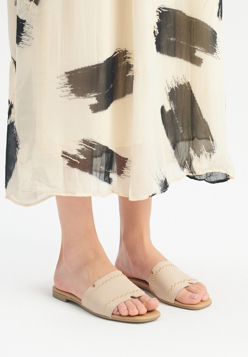 Beżowe klapki wykonane z gładkiej skóry, z ozdobnymi ćwiekami wzdłuż górnej krawędzi oraz otwartym noskiem.