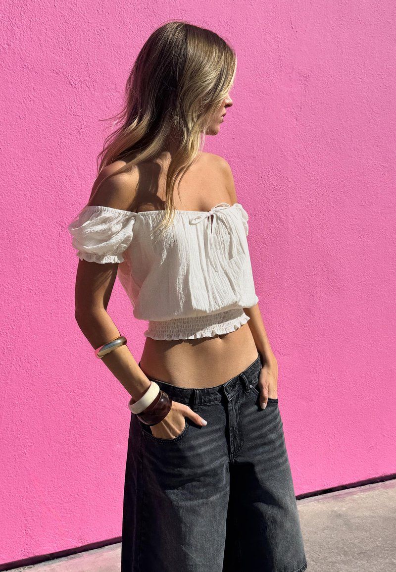 Mladá žena v bílém krátkém topu bez ramínek a černých širokých kalhotách stojí s rukama v kapse před jasně růžovou zdí.