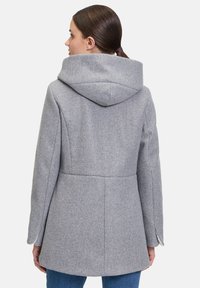Grau-wollner Mantel mit Kapuze, taillierter Schnitt und seitlichen Schlitzen. Der Stoff wirkt weich und strukturiert, was Wärme und Komfort verstärkt.