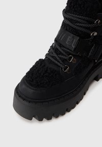Zwarte chunky laarzen met een textuur die lijkt op bont, een robuuste rubberen zool met diepe noppen, en metallic accenten op de veters en band.