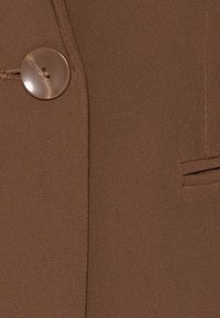 Tessuto marrone con finitura testurizzata, caratterizzato da un bottone rotondo e una tasca laterale, progettato con linee pulite e dettagli su misura.