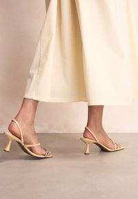 Sandales à talons beiges à brides avec une fine bride autour de la cheville et un petit talon courbé. Matière lisse avec un accent argenté brillant sur l'orteil.