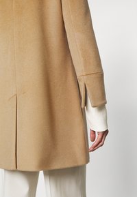 Manteau en laine couleur camel avec des fentes aux manches et au dos, porté par-dessus un pull blanc à manches longues et un pantalon blanc, montrant partiellement le dos et le bras.