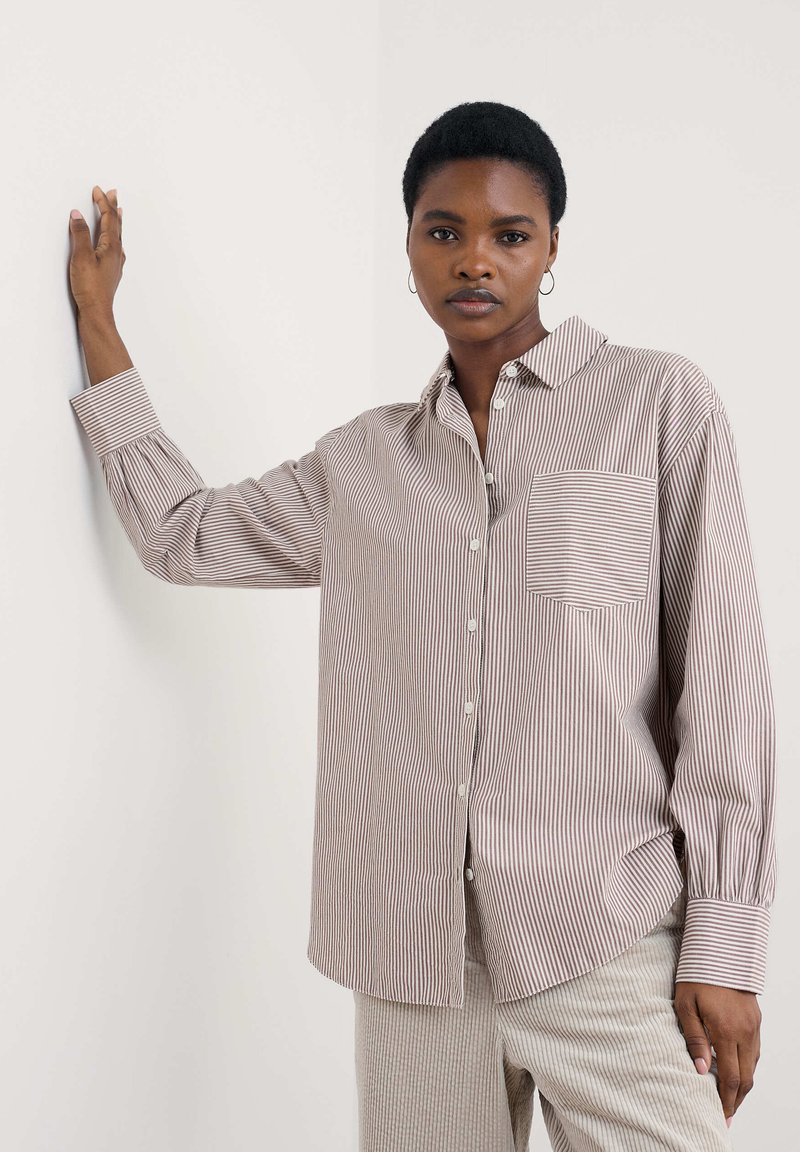 Camicia a righe in marrone chiaro e bianco, con colletto e taschino. Le maniche sono leggermente arricciate ai polsini.