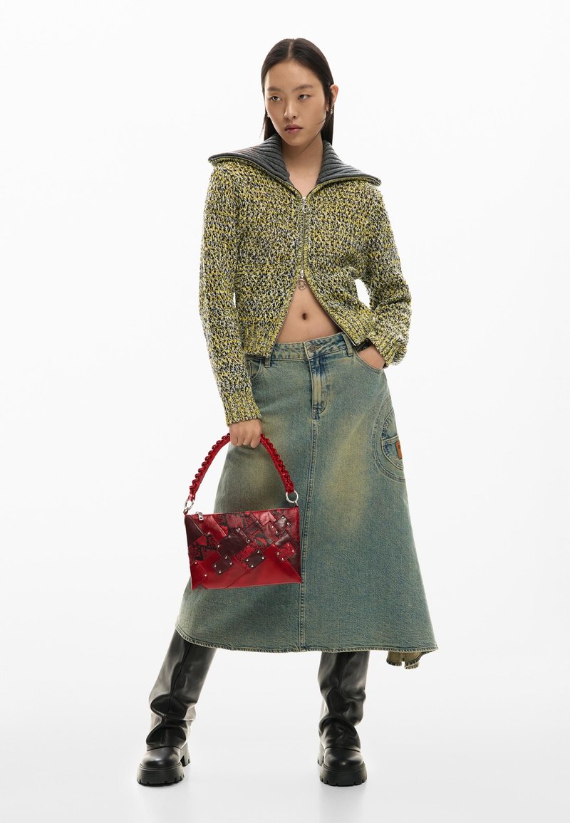 Woman wearing a cropped yellow-gray knit sweater, long denim skirt, black boots, holding a red handbag with a patterned handle on white background.