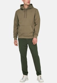 Sweat-shirt vert olive avec une poche avant, capuchon à cordon et poignets côtelés. Associé à un pantalon vert foncé et des baskets blanches.