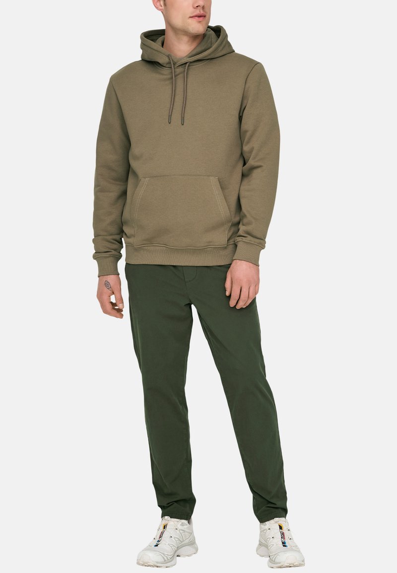 Sweat-shirt vert olive avec une poche avant, capuchon à cordon et poignets côtelés. Associé à un pantalon vert foncé et des baskets blanches.
