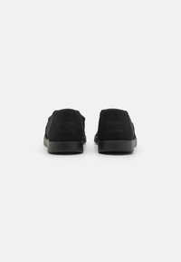 Black slip-on shoes with a textured fabric upper and flat rubber sole. Features minimal branding on the heel and a low-profile design.