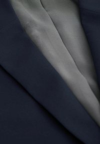 Blazer azul marino con una tela suave y texturizada y un forro gris claro que presenta un sutil patrón de rayas. El cuello abierto revela detalles interiores.