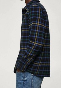 Camicia a scacchi di flanella in colori scuri, con linee blu, verdi e gialle, colletto con bottoni e maniche lunghe, abbinata a jeans di denim.
