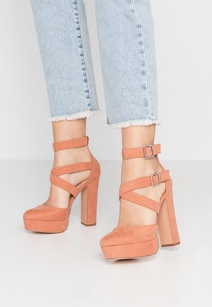 Peach suede platform heels with a chunky block heel, featuring crossed straps and silver buckles around the ankle.