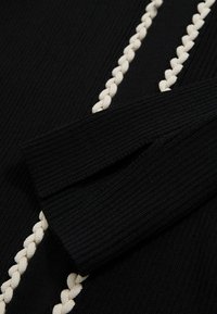 Közelkép egy fekete, texturált szöveten, amelyen krémszínű, fonott részlet található az varrás mentén, bal oldalon pedig egy hajtott ujj látható.