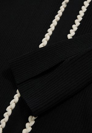 Nærbillede af et sort, struktureret stof med en flettet cremefarvet detalje langs sømmen, hvor en foldet ærme er synlig til venstre.