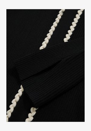 Nærbillede af et sort, struktureret stof med en flettet cremefarvet detalje langs sømmen, hvor en foldet ærme er synlig til venstre.