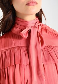 Rosa Seidenbluse mit hohem Kragen, versehen mit Rüsche-Details und einem gebundenen Gürtel. Angenehme Textur und fließendes Design mit plissierten Details.