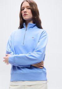 Γυναίκα με κοντά μαλλιά που φοράει ανοιχτό μπλε φούτερ με φερμουάρ και με σταυρωμένα χέρια μπροστά σε απλό λευκό φόντο.