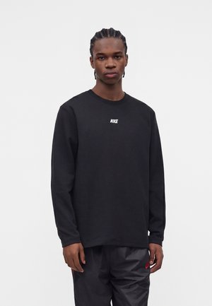 Junger Mann mit geflochtenem Haar, der ein schwarzes Nike-Langarmshirt und dunkle Hosen trägt, steht vor einem schlichten weißen Hintergrund.