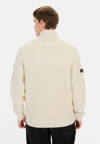 Kræmfarvet strikket sweater med høj krave, struktureret mønster og ribbede manchetter og kant. Har et logo-patch på den venstre ærme.