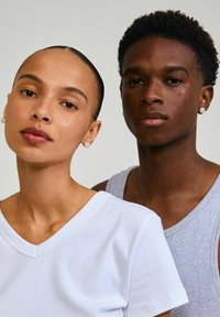 Due persone posano vicine. La persona a sinistra indossa una t-shirt bianca a V; la persona a destra ha un top grigio senza maniche.