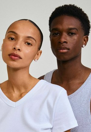 Due persone posano vicine. La persona a sinistra indossa una t-shirt bianca a V; la persona a destra ha un top grigio senza maniche.