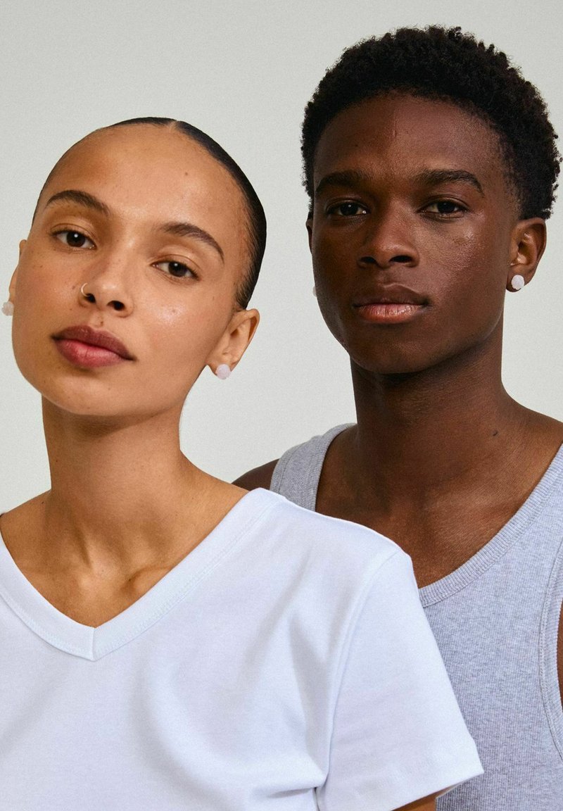Due persone posano vicine. La persona a sinistra indossa una t-shirt bianca a V; la persona a destra ha un top grigio senza maniche.