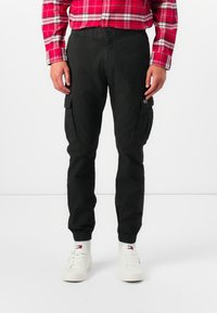 Pantalones cargo negros con cintura y puños elásticos. Presentan dos bolsillos laterales y una tela texturizada. Llevados con zapatillas blancas y una camisa de cuadros roja.