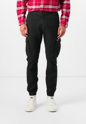 Pantalones cargo negros con cintura y puños elásticos. Presentan dos bolsillos laterales y una tela texturizada. Llevados con zapatillas blancas y una camisa de cuadros roja.