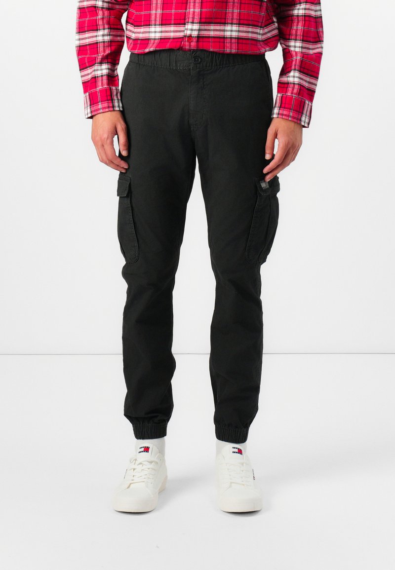 Pantalones cargo negros con cintura y puños elásticos. Presentan dos bolsillos laterales y una tela texturizada. Llevados con zapatillas blancas y una camisa de cuadros roja.