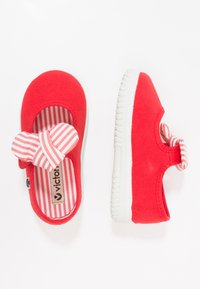 Par de zapatos rojos para niños pequeños con suelas blancas, que cuentan con un interior a rayas y un lazo de tela a rayas en la correa.