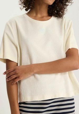 Femme portant un haut en maille crème à manches courtes avec un col rond et une jupe rayée bleu marine et blanc, les bras doucement croisés sur le torse.