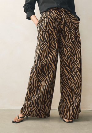 Pantalon à jambes larges avec rayures tigre marron et noire porté avec une chemise noire et des sandales à talons noires sur un sol gris clair.