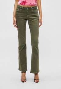 Πράσινο ελιάς, καμπανιστό τζιν με μπροστινές τσέπες, εμφανείς ραφές και κούμπωμα με κουμπί. Φορεμένο με πολύχρωμα σανδάλια με τακούνια.