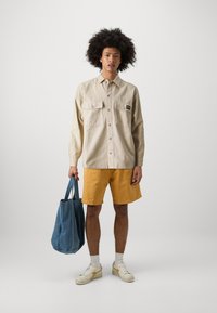 Beige button-up shirt with two front pockets, paired with yellow shorts. The model holds a blue tote bag and wears white sneakers. Smooth fabric.