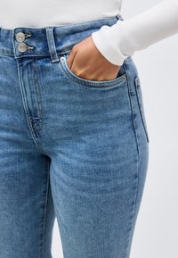 Χέρι στην τσέπη τζιν μεσαίου πλυσίματος σε μπλε χρώμα με διπλό κούμπωμα με κουμπιά, φορεμένο με λευκό μπλουζάκι μακριάς μανίκιας σε απλό φόντο.