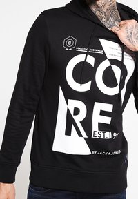 Sudadera negra con una gran gráfica blanca que incluye el texto "CORE EST. 1990" y formas geométricas. Tejido suave con capucha y cordón.