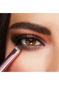En nærbilde av et øye med brun iris, lagdeltøyemakeup i rosa og svarte toner, og en sminkebørste med metallisk rosa håndtak som påfører skygge.