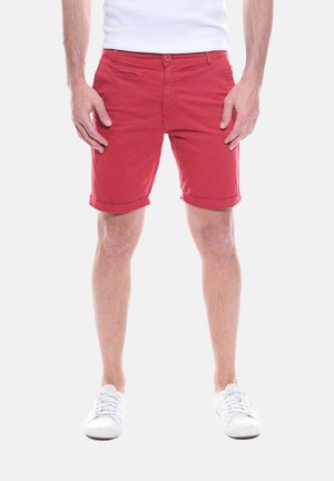 Homme portant un short rouge retroussé et des baskets blanches, debout avec les mains détendues le long du corps sur un fond uni.