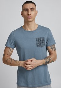 Blå bomulds T-shirt med rund halsudskæring, der har en sort mønstret brystlomme. Korte ærmer og afslappet pasform. Glat tekstur.