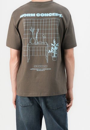 Personne portant un t-shirt marron avec un dessin au trait bleu d'une table, des plantes et le texte "NORM CONCEPT" ainsi que les coordonnées au dos.