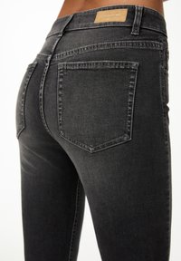 Jeans in denim nero con vita media, dotati di tasche posteriori e una superficie testurizzata. La cintura ha un'etichetta in pelle marrone chiaro.