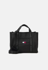 Musta kantokassi, jossa on tekstuuripintainen kangas, kaksi lyhyttä kantohihnaa ja irrotettava olkahihna. Koristeltu "TOMMY JEANS" -brändillä ja logo-patchilla.
