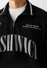 Svart zip-up pullover med hög krage, med grå text och ett vitt innerlager. Texten innehåller "Non-stop nattliv & livlig musikscen."