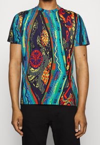 Man i en kortärmad, flerfärgad mönstrad stickad skjorta med abstrakta geometriska och vågiga mönster, tillsammans med svarta byxor.