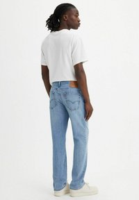 Jeans de mezclilla azul claro con un corte relajado, que cuentan con bolsillos traseros, un parche de cuero marrón y una camiseta blanca combinada con zapatos beige.