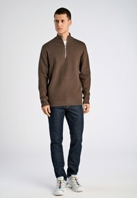 Brauner Zip-Pullover mit hohem Kragen, gestrickter Textur, kombiniert mit dunklen Jeans und weißen Sneakers mit farblichen Akzenten.