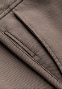 Gros plan sur un tissu marron avec des coutures détaillées et l'ouverture d'une poche passepoilée, montrant la texture et le savoir-faire en couture.