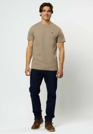 Noor mees seisab, kandes beežikat lühikeste varrukatega t-särki, tumelisi siniseid teksapükse ja pruune nahast paeltega saapaid, naeratades kaamerale.