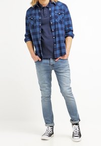 Casualny strój składający się z niebieskiej flanelowej koszuli w kratę noszonej na granatowym polo, jasnoniebieskich dżinsów i czarnych trampków za kostkę.