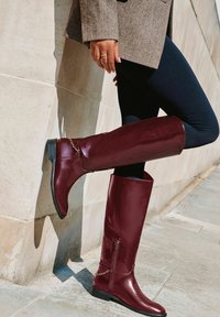 Burgundzkie kozaki za kolano wykonane z błyszczącej skóry, z bocznym zamkiem błyskawicznym i dekoracyjnym łańcuszkiem w okolicy kostki, z niskim obcasem.